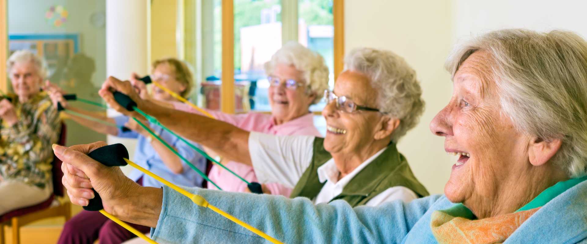 Large group of happy enthusiastic elderly ladies exercising in a gym sitting in chairs doing stretching exercises with rubber bands